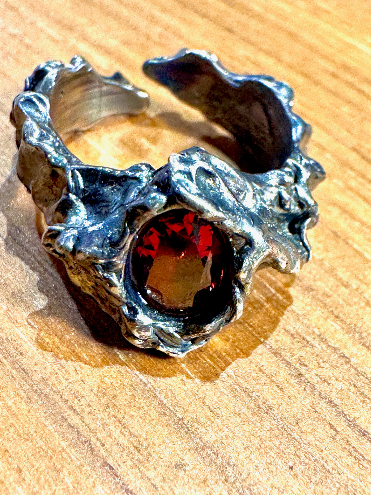 A rustic, handcrafted silver ring with a textured, organic shape and a central dark red gemstone, displayed on a wooden surface.